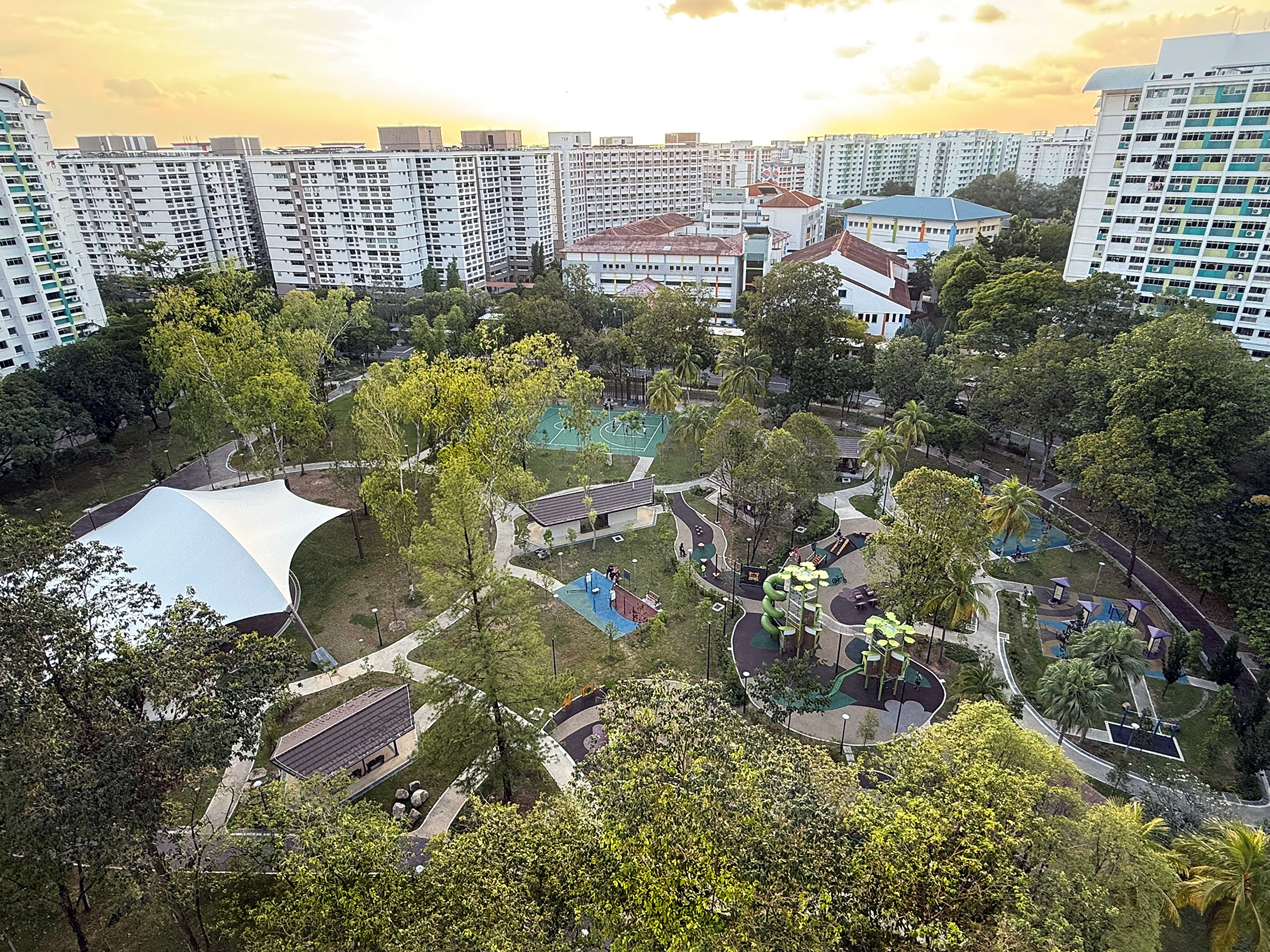 Aerial view of a tree-filled park with playgrounds and paths, surrounded by tall apartment blocks at sunset.